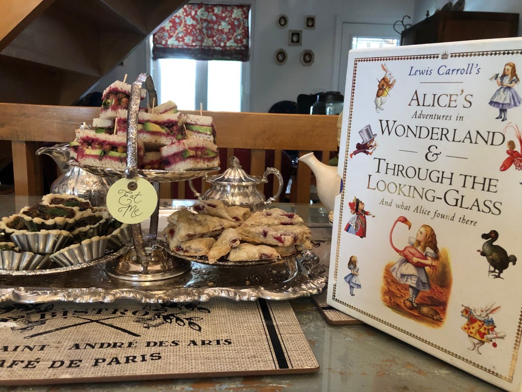 Lewis Carroll's Alice's Adventures in Wonderland & Through the Looking Glass and What Alice Found There, next to a display of tiny sandwiches, mini tarts, and scones. There's a silver sugar bowl and the edge of a teapot in the background. A little label that says "eat me" is on the display.