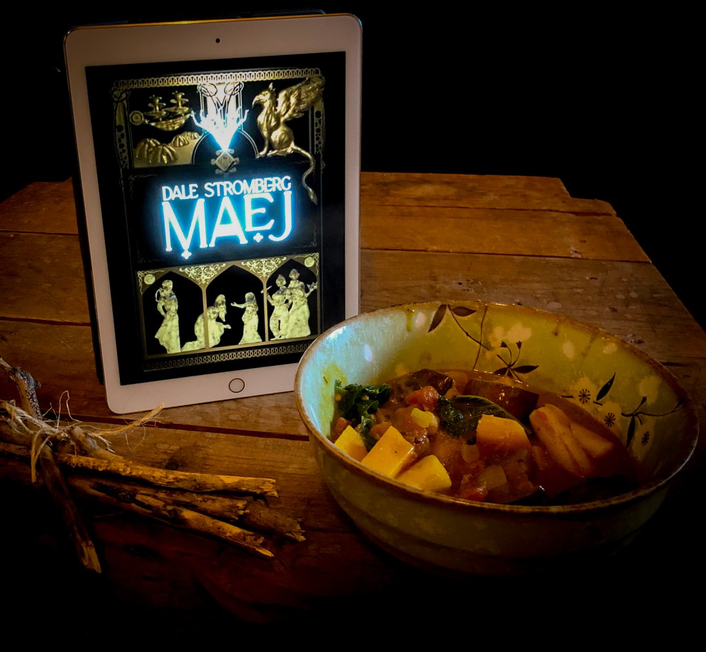 An ebook copy of Maej by Dale Stromberg on an iPad. It's sitting on a rustic wood coffee table. There's a bowl of vegetable stew and some sticks bundled together.