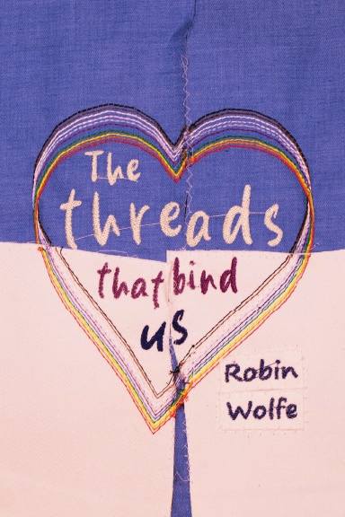 The Threads That Bind Us by Robin Wolfe. Image is an embroidered heart on two colours of fabric that divide the cover vertically with an uneven seam going down the middle. The heart is embroidered with multiple threads forming a rainbow.